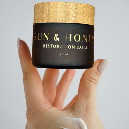 Hand holding a jar of 'Sun & Honey Tallow Restoration Balm' with a bamboo lid.