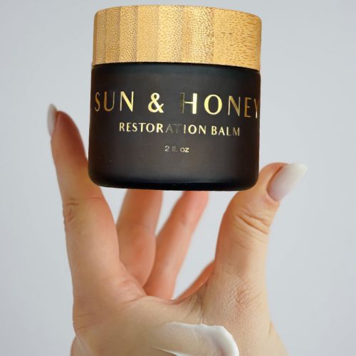Hand holding a jar of 'Sun & Honey Tallow Restoration Balm' with a bamboo lid.